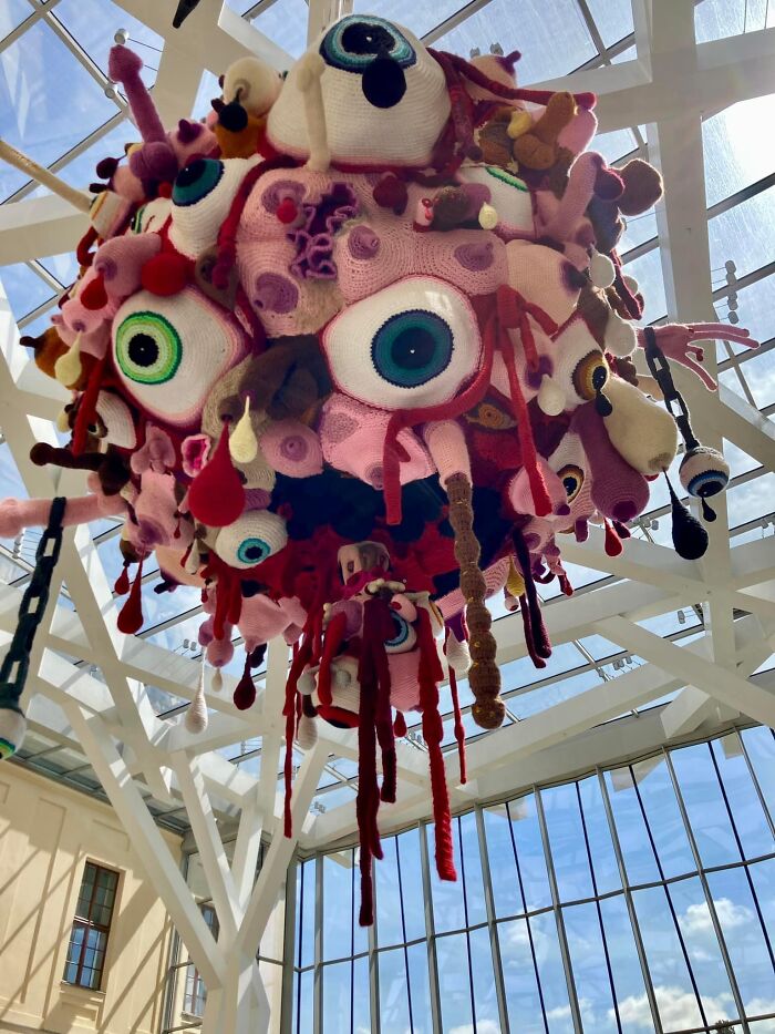 Crocheted art installation with multiple colorful eyeballs hanging from a ceiling, showcasing DIY crafts in a museum setting.