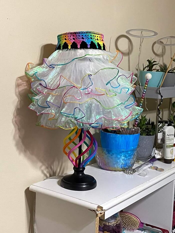 Horrendous DIY lamp with multicolored swirl base and frilly, rainbow-trimmed shade on a white cabinet.