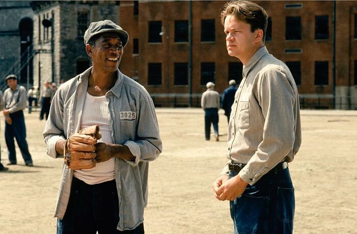 Two men in casual 90s attire standing outdoors at a prison yard, surrounded by other men in similar outfits.