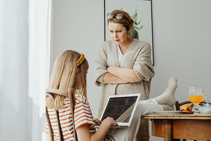 Teen girl using laptop, adult woman standing nearby looking concerned, highlighting teen's choice to go out instead of babysitting.