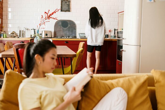 Roommate reads on sofa while someone washes dishes in messy apartment kitchen.