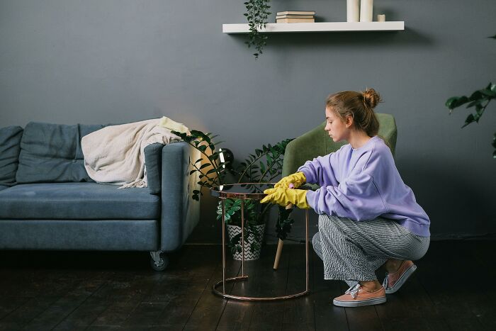 College student cleaning messy apartment living room wearing yellow gloves, next to a couch and plants.