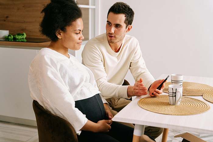 A pregnant woman talking to a man over coffee at a table, holding a mobile phone.