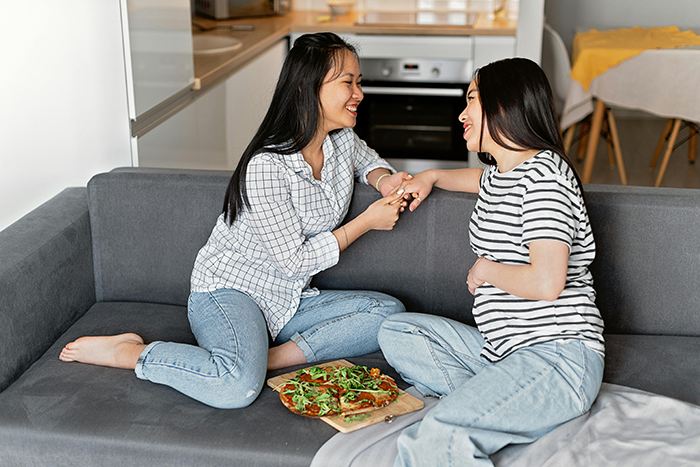 Two women on a sofa share a joyful moment, holding hands, with a pizza beside them.