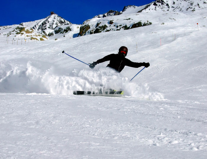 Skier on snowy trail with mountains in the background, wearing black gear, mid-turn, creating snow spray.
