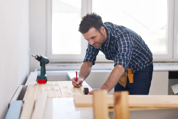 Person crafting a wooden item, wearing a plaid shirt; a drill and measuring tools are visible on the table.