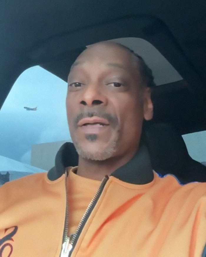 Snoop Dogg in an orange jacket sitting in a car, with a plane visible in the background sky.