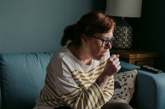 Person sitting on a couch, wearing glasses and a striped shirt, coughing, with a lamp and remote visible nearby.