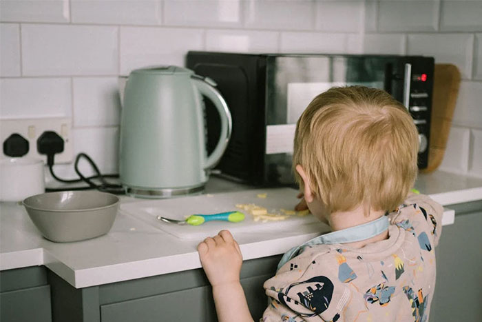 A child in a kitchen reaching for food, with a kettle and microwave in the background.