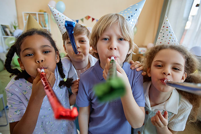 Children at a party wearing hats, blowing noisemakers, with siblings and no presents visible.