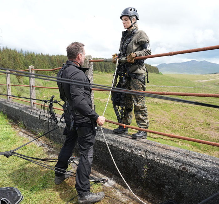 Special Forces Season 3 stars including Ali Fedotowsky-Manno in rappelling gear on a ledge during training in a mountainous setting.