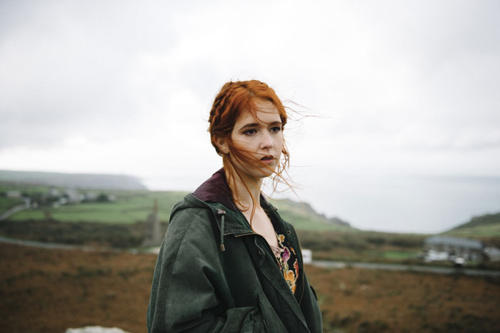 Woman with red hair standing outdoors, wearing a green jacket, with a scenic landscape in the background.