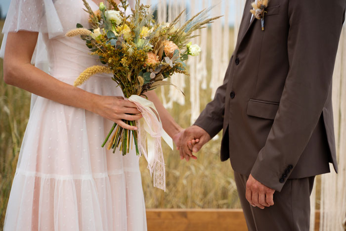 Bride holding bouquet beside groom in brown suit at outdoor wedding ceremony.