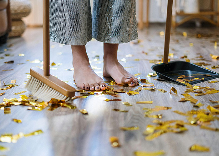 Barefoot person sweeping gold confetti off a wooden floor into a dustpan, illustrating a hotel housekeeper's unusual find.