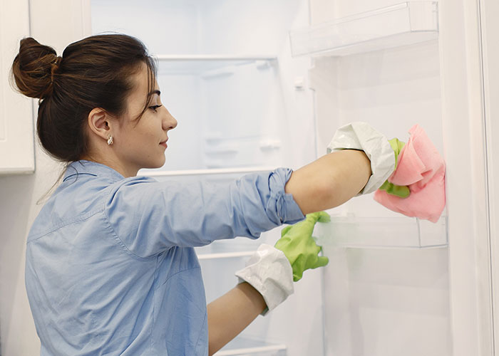 Hotel housekeeper cleaning a fridge in a guest room, wearing green gloves and using a pink cloth.