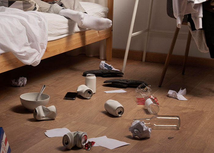 Messy hotel room with crumpled cans, scattered papers, and a bowl on the floor, seen by housekeepers.