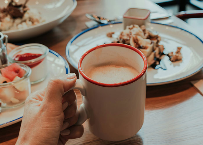 A hand holding a coffee mug at a breakfast table, featuring pancakes and fruit.