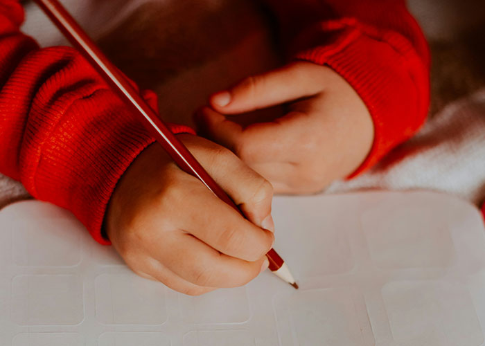 Child writing with a pencil on paper, wearing a red sweater, related to creative punishments.