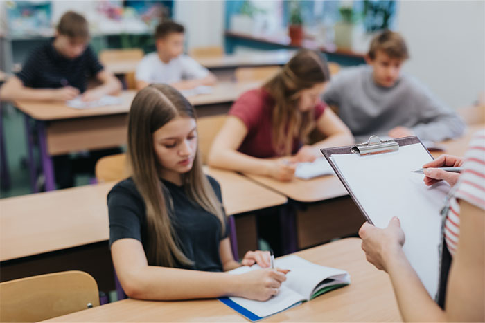 Student sits in class with a focused expression as teacher holds clipboard, reflecting on name pronunciation incident.