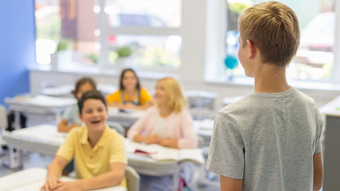 Student stands in classroom, classmates smiling, capturing a priceless look.