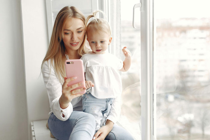 Woman with child looking at phone by a window, related to private home tour themes. Woman with child looking at phone by a window, related to private home tour themes.