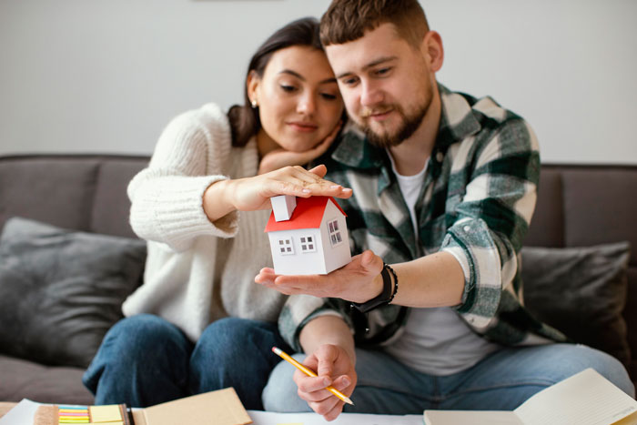 Couple holding a model house, appearing thoughtful during a home tour. Couple holding a model house, appearing thoughtful during a home tour.