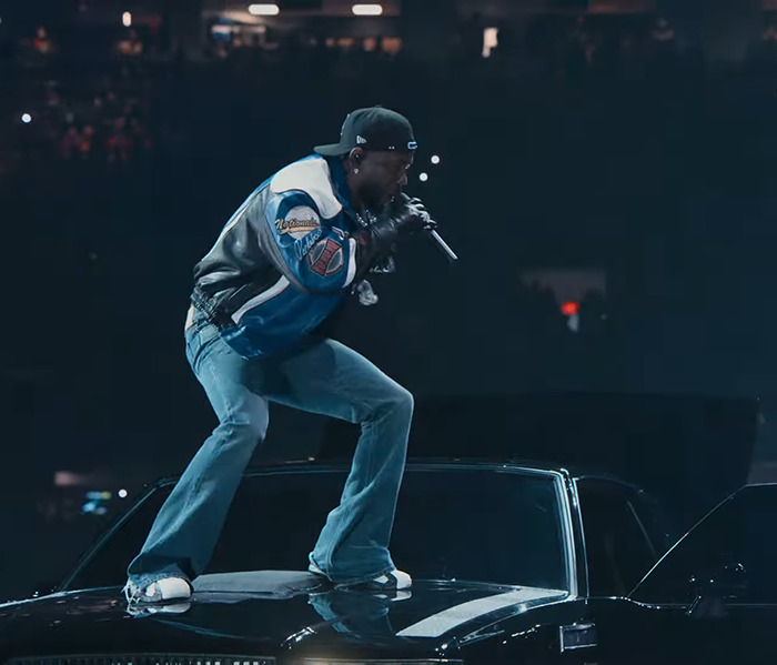 Kendrick Lamar. performing on a stage, wearing a cap and jacket, under dramatic lighting.
