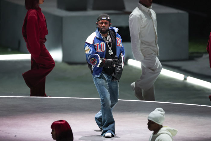 Kendrick Lamar performing on stage during a halftime show, wearing a blue jacket and jeans. Kendrick Lamar performing on stage during a halftime show, wearing a blue jacket and jeans.