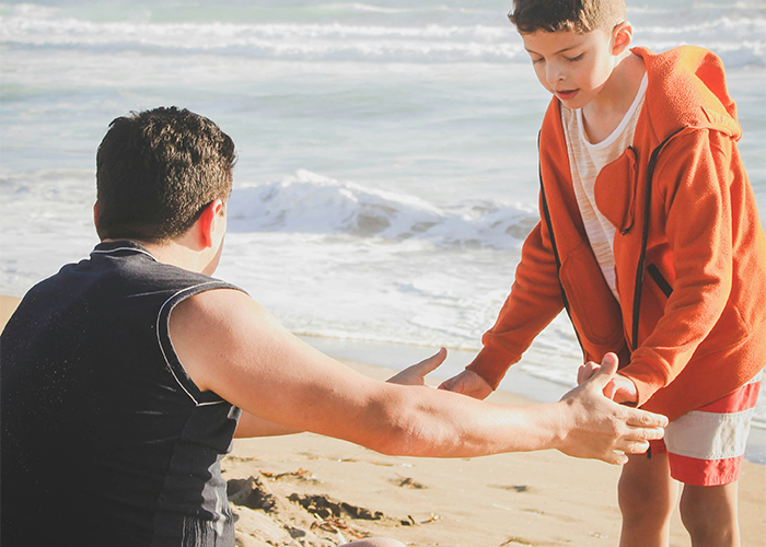 Man and boy in an orange hoodie at the beach, emphasizing support and family bonds.