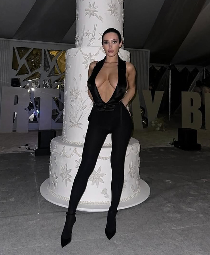 Bianca Censori in a revealing black outfit stands confidently in front of a large decorative cake.