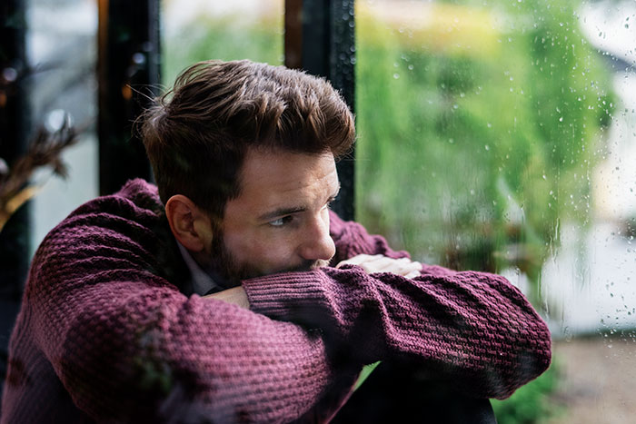 Man gazing thoughtfully out a rainy window, wearing a maroon sweater, symbolizing introspection about surprising things women learn.