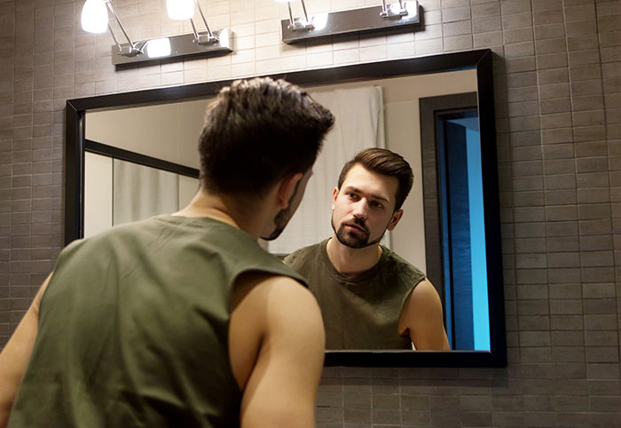 Man in a sleeveless shirt looking at his reflection in a bathroom mirror.