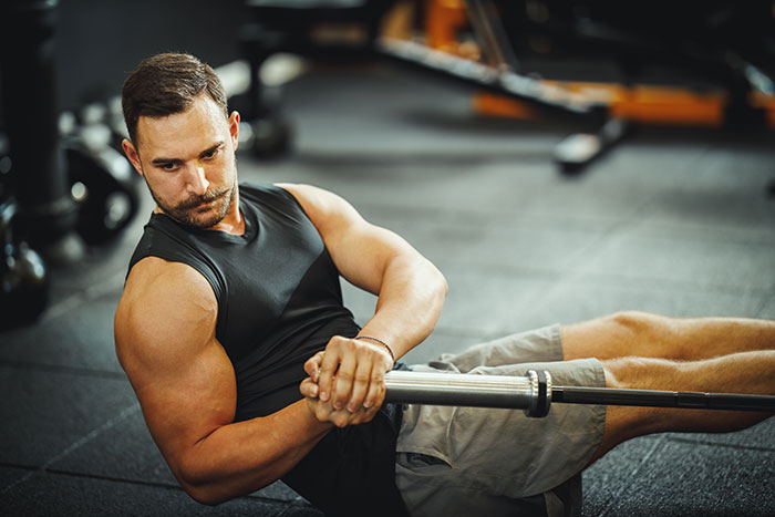 Man exercising at the gym in a black tank top, performing a seated twist with a barbell.