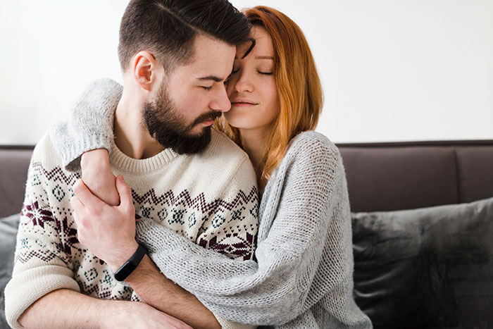 Couple embracing on a couch, woman with closed eyes and man wearing a patterned sweater.