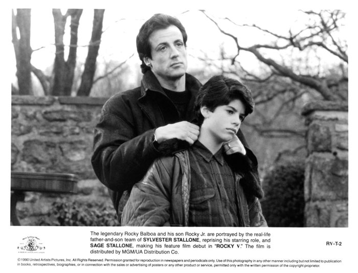 Sylvester Stallone with a young boy in a contemplative scene from a movie, both wearing dark jackets in an outdoor setting.