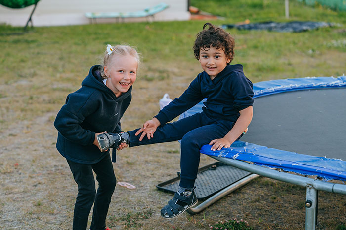 Two kids smiling while playing on a trampoline outdoors, wearing dark hoodies.