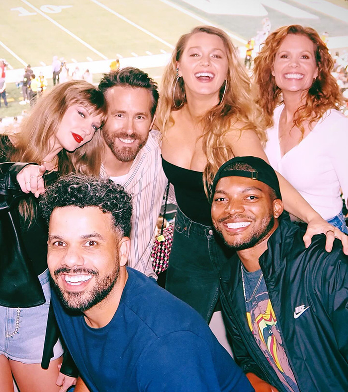 Taylor Swift, Blake Lively, and Ryan Reynolds poses with friends at a stadium, capturing a joyful moment in a lively atmosphere.