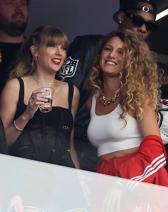 Taylor Swift and Blake Lively smiling at an event, one holding a drink, with NFL logo in the background.