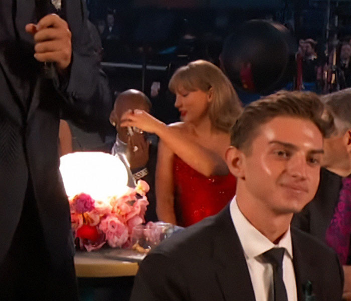 Young man in a suit at the 2025 Grammy Awards, with a woman in red reaching over a table adorned with flowers behind him.