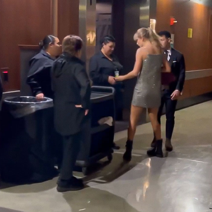 Taylor Swift handing cash tips to staff in a hallway at the Grammys, wearing a sparkly dress and boots.