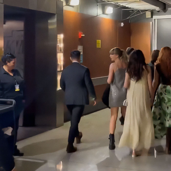 Taylor Swift leaving an event, wearing a silver dress, surrounded by people in an indoor hallway.