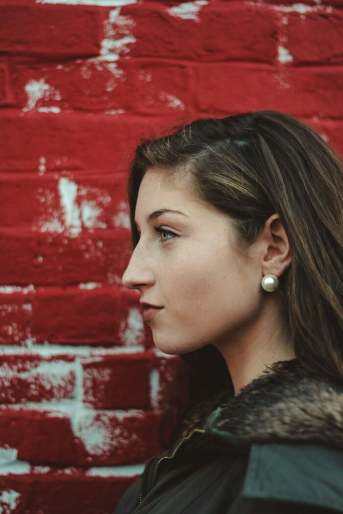 Profile of a woman with a prominent nose, wearing a pearl earring, in front of a red brick wall.
