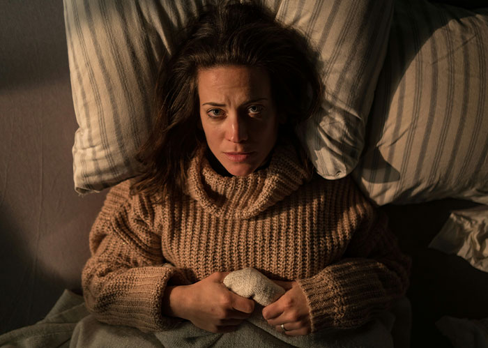 A woman lying in bed, looking concerned, wearing a brown sweater, surrounded by pillows. A woman lying in bed, looking concerned, wearing a brown sweater, surrounded by pillows.