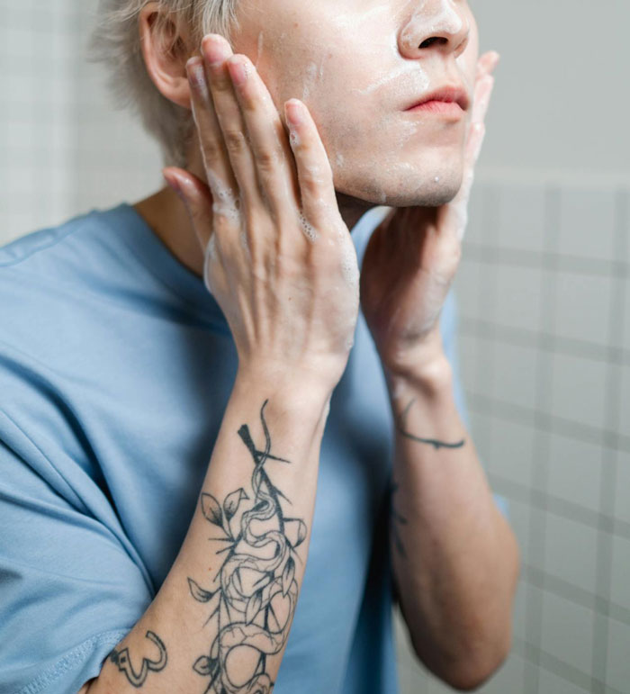 Teen applying skincare product, with tattoos visible on arms, in a tiled bathroom.