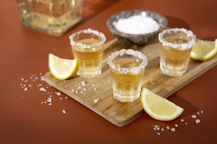 Three tequila shots with salted rims on a wooden board, surrounded by lemon wedges.