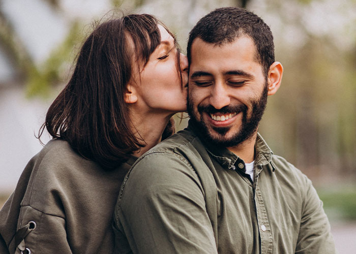 A woman affectionately kisses a smiling man on the cheek outdoors.