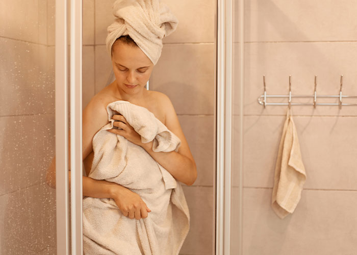 Woman wrapped in towel with towel on her head, standing in a shower.