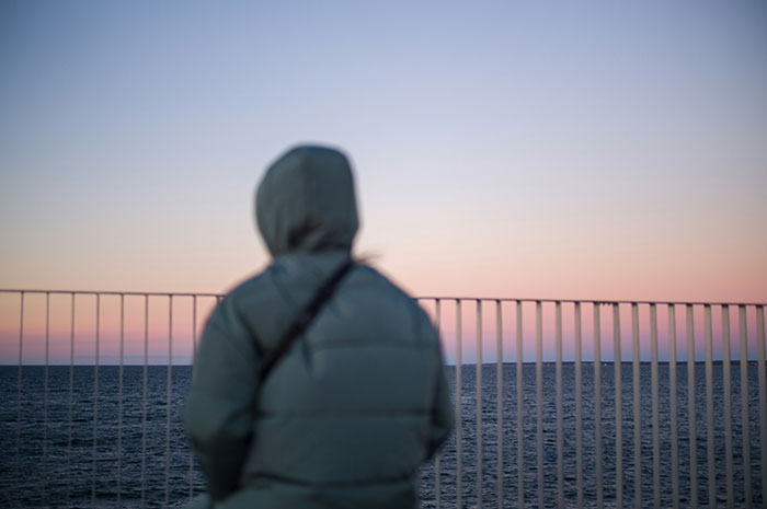 Person in a hooded jacket gazing at the ocean during sunset, capturing a moment of grief and reflection.