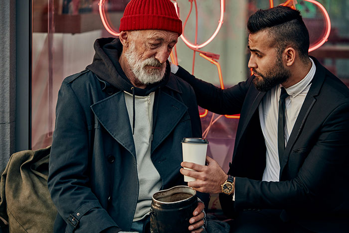 A younger man comforts an older man in a red hat, seated, both experiencing a moment of grief.