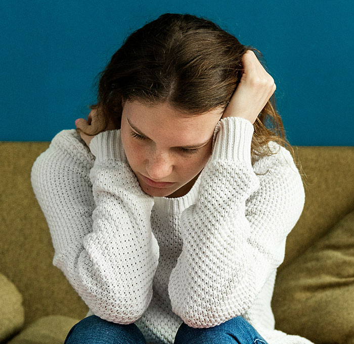 A woman in a white sweater expressing grief, sitting on a sofa with a blue wall background.